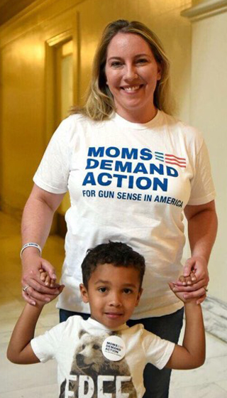 Woman wearing Moms Demand Action t-shirt holds the hands of a small boy in front of her.
